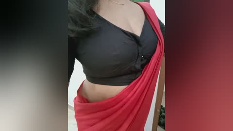 Media: Video of a woman wearing a black V-neck blouse and a red saree draped over her left shoulder. Her long, black hair falls over her right shoulder. Background is blurred.