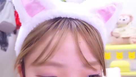 Media: A close-up video of a person with light brown hair, wearing a white cat ear headband, indoors. The background features a stuffed teddy bear and yellow kitchen items.