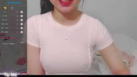 Media: Video of a smiling Asian woman with light skin, wearing a tight white t-shirt that shows her ample cleavage and nipples. Background includes a white bed with a pink pillow.