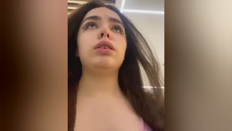 Media: Video of a young woman with long, dark hair and light skin, wearing a pink top, looking upwards with a neutral expression. The background features blurred beige walls and a ceiling.