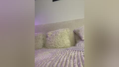 Media: A video of a cozy, dimly lit bedroom with a cream-colored bedspread featuring a textured, fluffy pillow. The room is softly illuminated by a purple light, creating a serene ambiance.