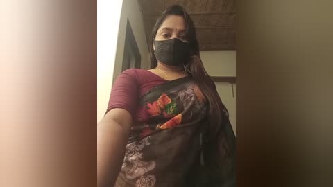 Media: Video of a South Asian woman with long black hair, wearing a black mask, maroon top, and floral sari, standing indoors with wooden ceiling beams in the background.