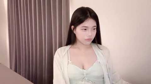 Media: Video of an Asian woman with long black hair, wearing a light blue lace top and a white cardigan, standing in front of vertical gray curtains against a beige wall.