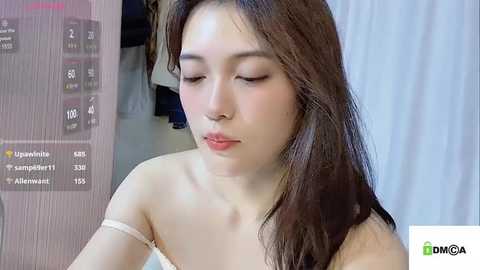 Media: A video of a young Asian woman with long brown hair, wearing a white strapless top, looking down with a contemplative expression. Background features a blue curtain and a partially visible wooden door.