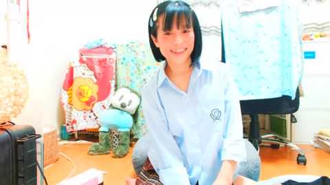 Media: Video of a smiling Asian woman with short black hair in a light blue button-down shirt, sitting at a desk with a wicker basket, stuffed animals, and colorful fabric on a wooden floor.