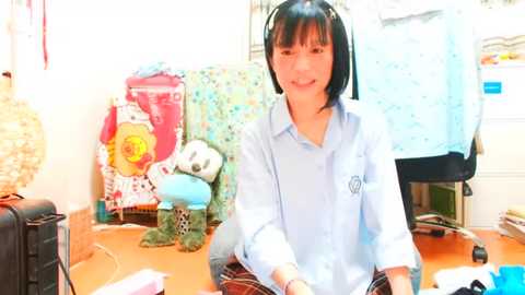 Media: Video of a smiling East Asian woman with shoulder-length black hair, wearing a light blue collared shirt and a plaid skirt, sitting at a cluttered desk with stuffed animals and colorful toys in a cozy, well-lit room.