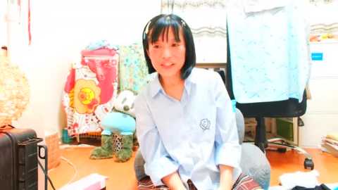 Media: Video of a young woman with straight black hair, smiling, seated at a cluttered desk with a blue shirt, surrounded by a sewing room with fabric, sewing machine, and wicker baskets.