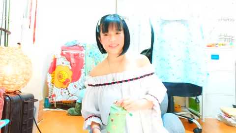 Media: Video of an Asian woman with short black hair and fair skin, wearing an off-shoulder white top, seated at a cluttered desk with colorful decorations.