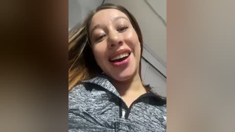 Media: Video of a smiling woman with light skin and shoulder-length brown hair, wearing a gray and black zip-up hoodie, in an indoor setting with white ceiling tiles.