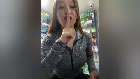 Media: Video of a woman with light skin and long brown hair, wearing a gray zip-up jacket, making a shushing gesture in a brightly lit convenience store filled with colorful shelves of snacks and drinks.