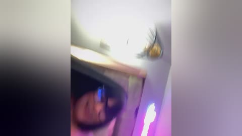 Media: A blurred video captures a young person with dark hair and glasses, sitting in a dimly lit room with a pink and purple light reflecting off the ceiling. The image is slightly out of focus.