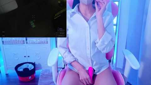 Media: Video of a light-skinned, slender woman wearing an open white button-up shirt, sitting on a pink gaming chair in a brightly lit room. She holds a pink dildo against her bare genitals.