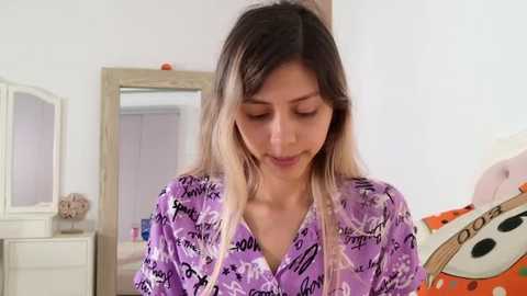 Media: Video of a young woman with long, dark blonde hair wearing a purple shirt with white text, standing in a bedroom with white walls, a mirror, and colorful pillows.