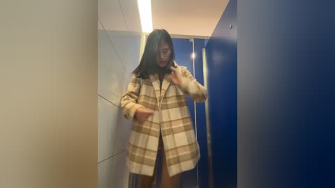 Media: Video of a young Asian woman with shoulder-length black hair in a narrow, tiled hallway, wearing a beige plaid coat and adjusting her hair, surrounded by blue and white walls.