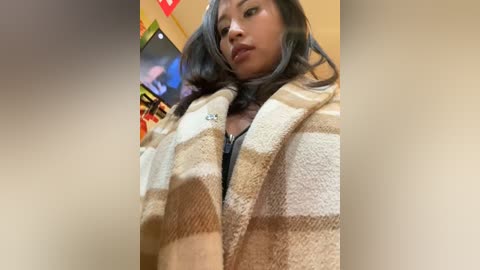 Media: Video of a young Asian woman with long, wavy black hair, wearing a beige and white striped blanket over a black top. Background shows a TV and colorful decor.