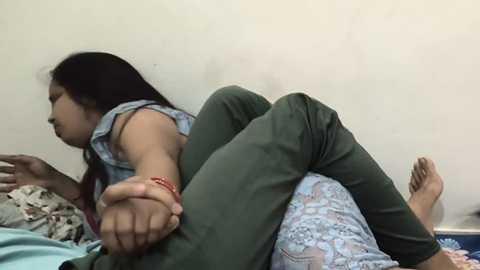 Media: Video of a young woman with long black hair, wearing a green shirt and floral pajama pants, lying on a bed, reaching out to another person, who is not visible.