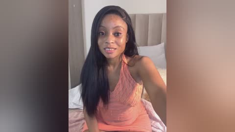 Media: Video of a young Black woman with medium-dark skin, straight black hair, and a slender build, wearing a pink halter dress, sitting on a bed with white sheets and a beige headboard in a minimalist bedroom.