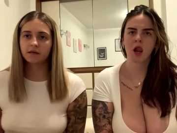 Media: Video of two young women with straight, long blonde hair, in white tops, one with tattoos and large breasts, one with small breasts, standing indoors, looking at each other.