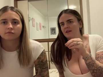 Media: Video of two young women with long hair, one with light brown hair and a white t-shirt, the other with dark hair, tattoos, and a white top. Both have a neutral expression. Background features a bathroom mirror and wall decor.