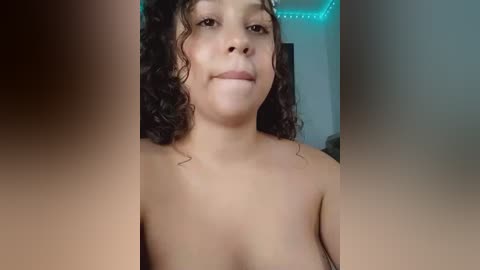 Media: Video of a topless young woman with curly hair, light brown skin, and a neutral expression, standing in a dimly lit room with green LED lighting.