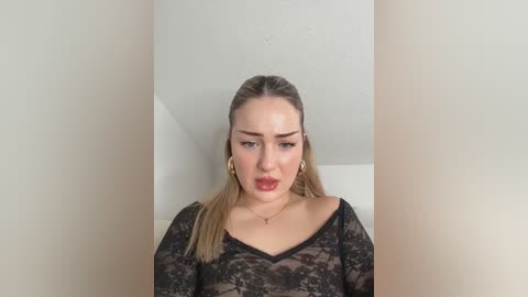 Media: Video of a fair-skinned woman with straight, shoulder-length blonde hair, wearing a black lace top, and a delicate gold necklace. She has a serious expression. Background is a plain white wall.