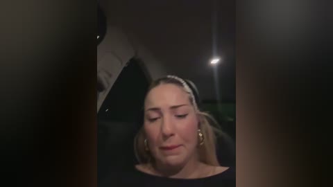 Media: A video of a woman with light skin and blonde hair, wearing a black top, sitting in a car with dim lighting, eyes closed, possibly sleeping.