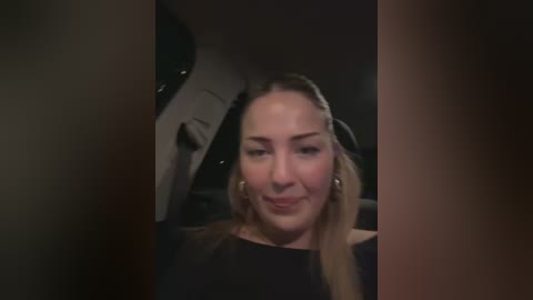 Media: A video of a young woman with fair skin and blonde hair, wearing a black top, smiling inside a dimly lit car.