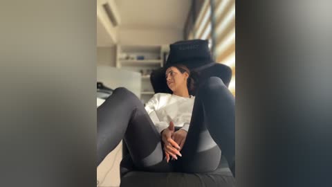 Media: Video of a young woman with light brown skin, wearing a white shirt and black tights, reclining on a black gaming chair in a brightly lit room with sunlight streaming through horizontal blinds.