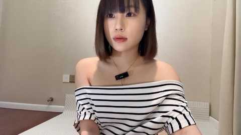 Media: A video of an East Asian woman with straight, shoulder-length brown hair and bangs, wearing a black and white striped off-shoulder top. She has a slender figure and is indoors against a plain beige wall.