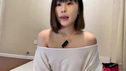 Media: Video of an Asian woman with shoulder-length straight brown hair, wearing a white off-shoulder top, standing indoors with beige walls and a wooden floor.