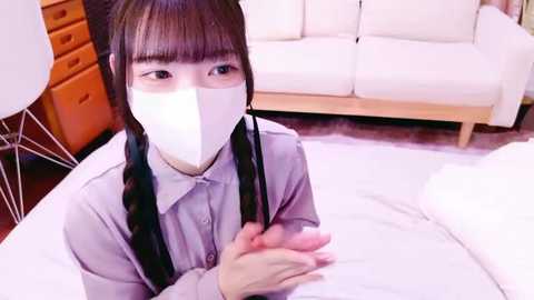 Media: Video of an Asian woman in a white mask, wearing a light gray shirt with black braids, seated on a bed, in a room with a white couch and wooden dresser.