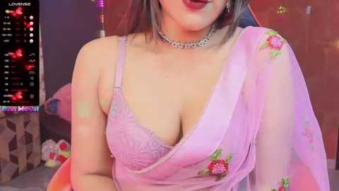 Media: A video of a woman in a pink saree with floral embroidery, wearing a revealing lace bra, and a silver choker. Background includes a live stream interface and a soft, colorful setting.