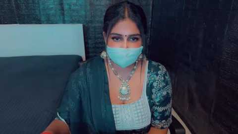 Media: Video of a South Asian woman with dark hair, wearing a traditional saree, green mask, and ornate jewelry, seated on a bed against a dark brick wall.