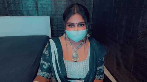 Media: Video of a woman in traditional South Indian attire, wearing a green saree with intricate embroidery, a silver necklace, and a face mask, seated on a bed with dark curtains in the background.