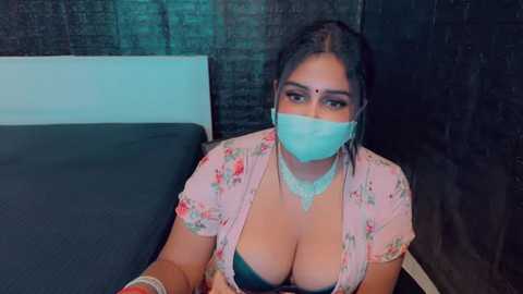 Media: Video of a South Asian woman with dark hair, fair skin, wearing a floral blouse, revealing cleavage, blue face mask, and gold necklace, sitting on a bed with dark curtains in the background.