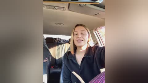 Media: Video of a smiling, light-skinned woman with shoulder-length blonde hair wearing a black zip-up hoodie, sitting in a car with beige seats and visible steering wheel.