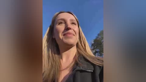 Media: A video of a smiling, fair-skinned, blonde woman wearing a black jacket, set against a clear blue sky with green trees in the background.