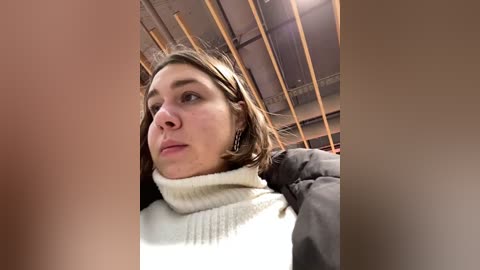 Media: Video of a young woman with fair skin and brown hair, wearing a white turtleneck sweater, looking thoughtful in an industrial, dimly lit space with exposed wooden beams.