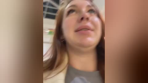 Media: Video of a Caucasian woman with fair skin, straight brown hair, wearing a gray top, smiling slightly, standing in a blurred indoor setting.