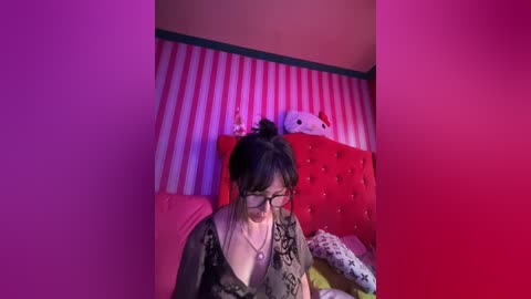 Media: A video of a woman with black hair in a bun, glasses, and a green dress, sitting on a bed with pink and white striped wallpaper, a red tufted headboard, and a plush Hello Kitty.