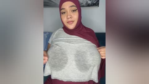 Media: A video of a South Asian woman with a medium complexion, wearing a maroon hijab and gray top, revealing her large breasts. She has arched eyebrows and full lips.