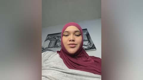 Media: Video of a light-skinned woman with a closed-mouth smile, wearing a maroon hijab and a grey and white top. Background features a monochrome painting of a classical building.