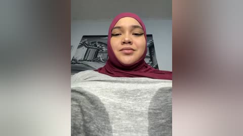 Media: Video of a young woman with light brown skin, wearing a red hijab and grey top, sitting indoors with a black-and-white architectural print on the wall behind her.