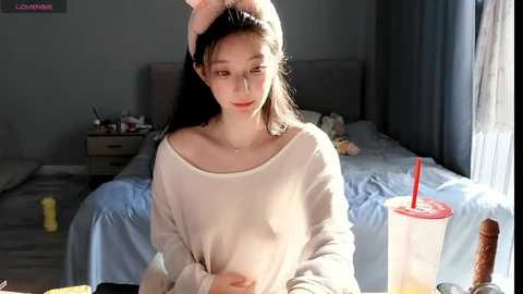 Media: Video of a young Asian woman with long black hair, wearing a loose white sweater, sitting on a bed with a messy room background.