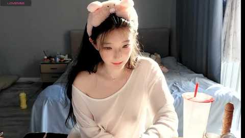 Media: A video of an Asian woman in a white off-shoulder sweater with a pink bunny headband, sitting by a window in a cozy bedroom.