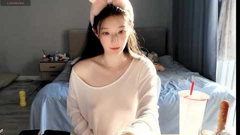 Media: Video of a young Asian woman with long black hair, wearing a white off-shoulder sweater, sitting on a bed in a dimly lit bedroom.