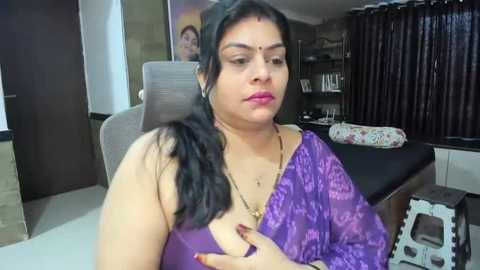 Media: Video of a South Asian woman with medium skin tone, wearing a purple saree and bindi, seated in a modern living room with wooden furniture and dark curtains.