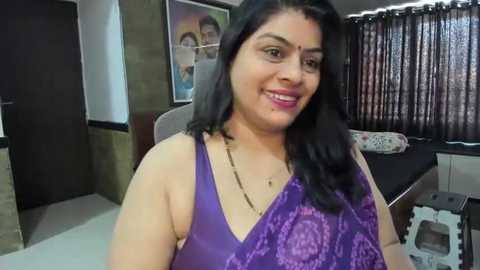 Media: Video of a smiling Indian woman with long black hair, wearing a purple saree with floral patterns, in a modern living room with framed family photos and wooden blinds.