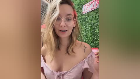 Media: Video of a Caucasian woman with blonde hair, wearing glasses, a pink off-shoulder top, and a tattoo on her left shoulder, standing in front of a green wall with a red \"Kitty\" sign.