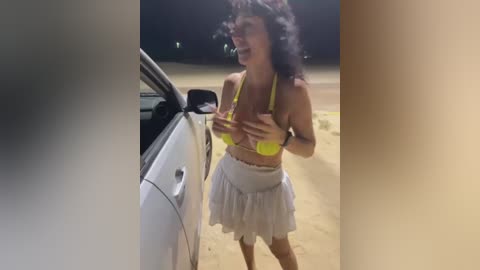 Media: Video of a curvy woman with dark hair, wearing a yellow bikini top and white skirt, laughing while leaning on a white car, on a sandy beach at night.